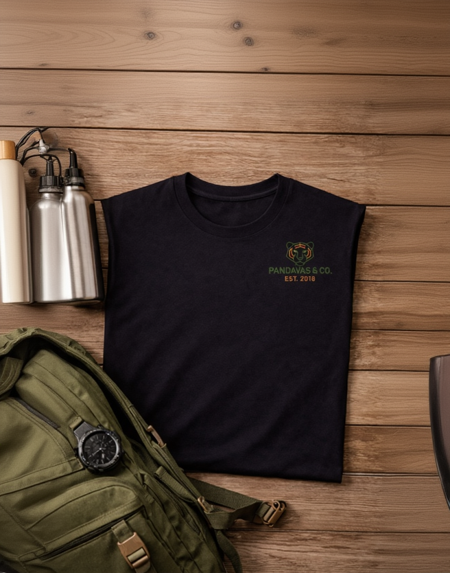 Black t-shirt with a logo and text on a wooden surface next to a green backpack and silver water bottle.