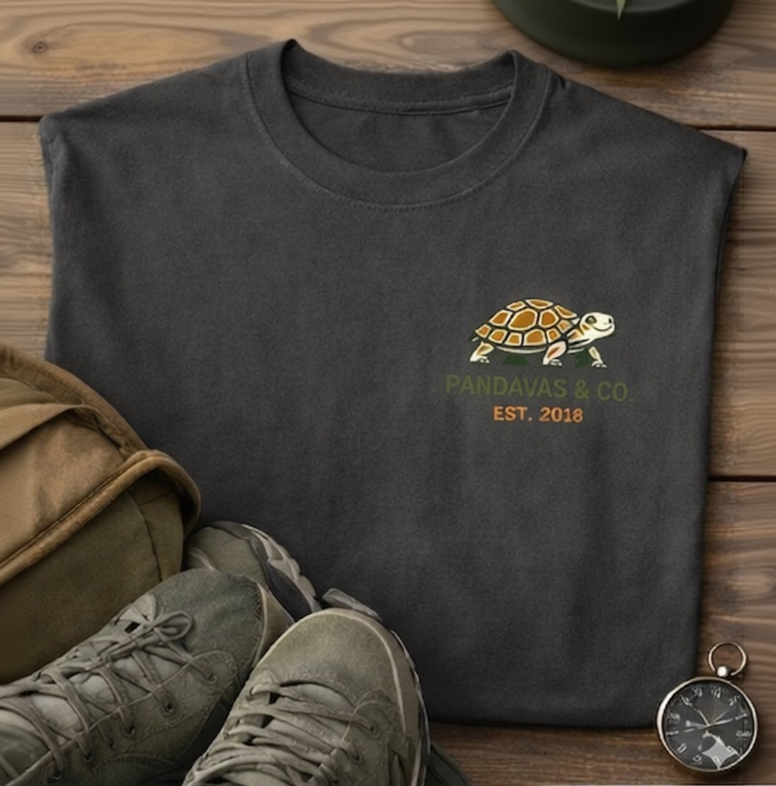 Dark gray t-shirt with turtle logo and 'Pandavas & Co.' text, paired with green shoes and a bag on a wooden surface.