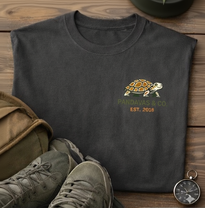 Dark gray t-shirt with turtle logo and 'Pandavas & Co.' text, paired with green shoes and a bag on a wooden surface.